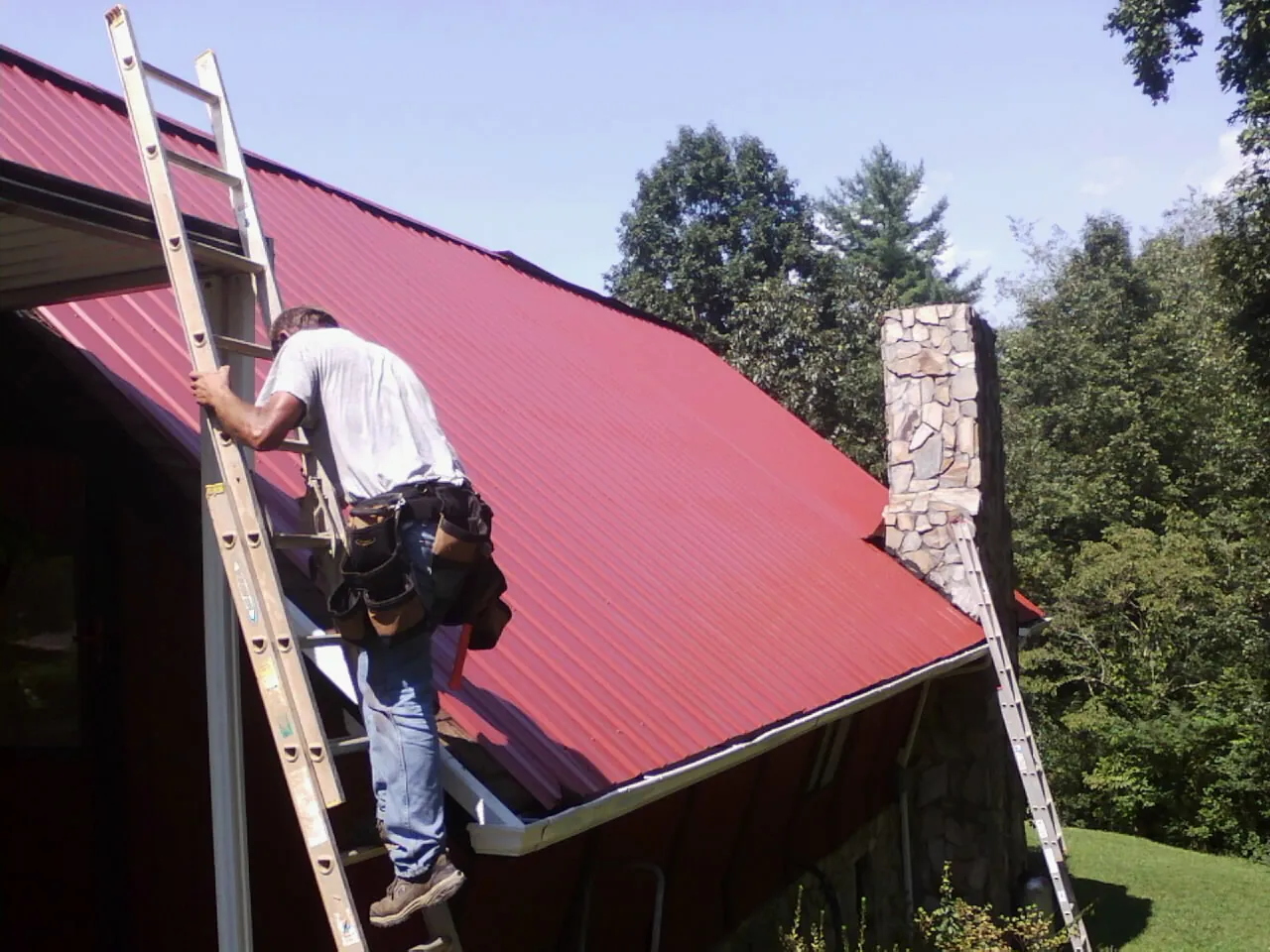 Roofer on a ladder beside a red metal roof and stone chimney — Soffit & Fascia Repair by Roofing Brothers in Bridgeport